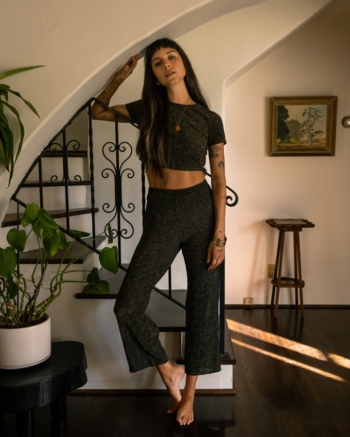 A woman standing in front of a staircase in a black crop top and the Dandy Del Mar Hydra Sheer Shimmer Pant - Onyx with elastic waist for a shimmer effect.