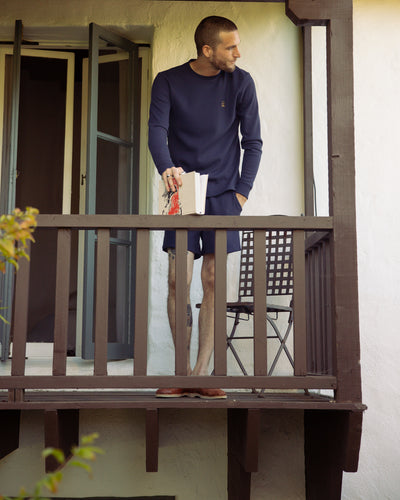 A man in the Dandy Del Mar Cannes LS Waffle Knit Shirt in Luxe Navy and shorts stands barefoot on a small balcony, holding a book and looking left. A chair sits behind him near the open door.