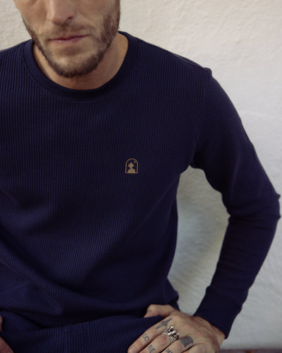 A man in Dandy Del Mar’s The Cannes LS Waffle Knit Shirt in Luxe Navy, featuring a small chest logo, sits against a light wall wearing several rings.