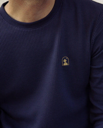 A person wearing Dandy Del Mar’s The Cannes LS Waffle Knit Shirt in Luxe Navy, featuring textured fabric and a small yellow embroidered chess piece with star logo on the chest.