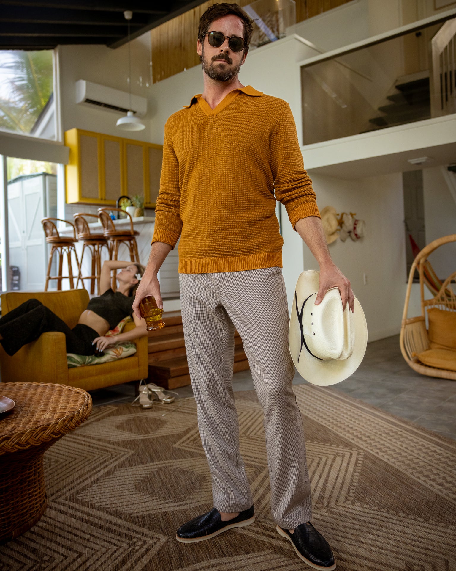 A man indoors wears Dandy Del Mar's "The Sebastian Long Sleeve Polo" in Burnt Sienna with gray pants, holding a hat and drink. He sports sunglasses, while a woman lounges on a couch amid mid-century modern decor.