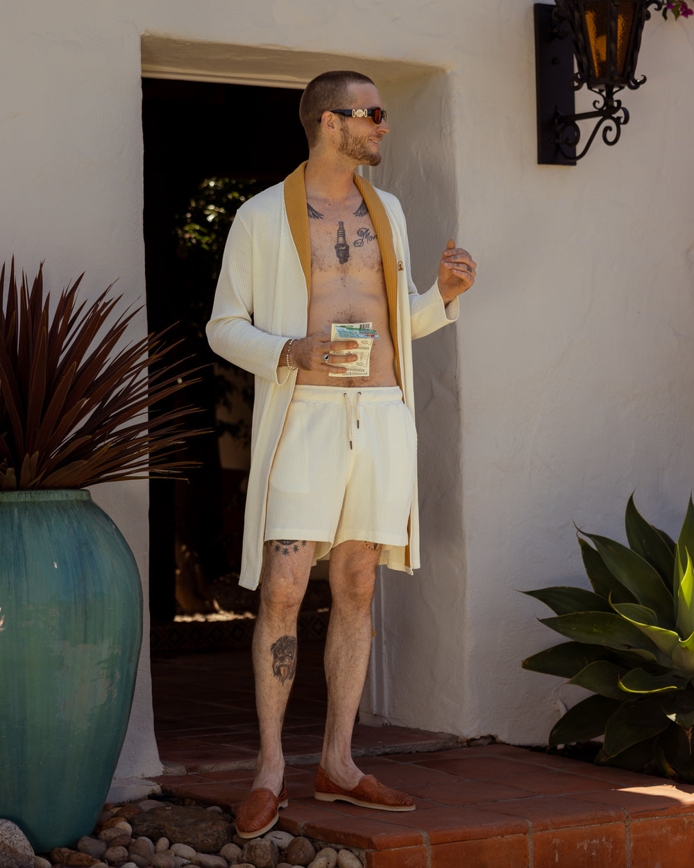 A man in a white shirt and shorts standing in front of a house wearing The Cannes Waffle Knit Robe - Vintage Ivory by Dandy Del Mar.