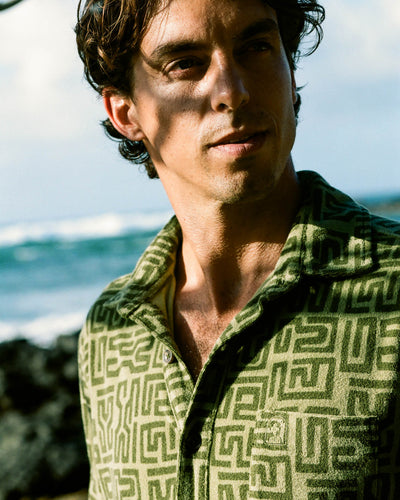 A man in the Dandy Del Mar Tropez Terry Cloth Shirt – Berdea Tayana Print stands outdoors by the ocean on the Côte d'Azur, sunlight and shadows playing across his face.