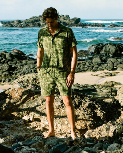 A man in Dandy Del Mar’s Tropez Terry Cloth Shirt - Berdea Tayana Print with matching shorts stands barefoot on rocky terrain by the Côte d'Azur.