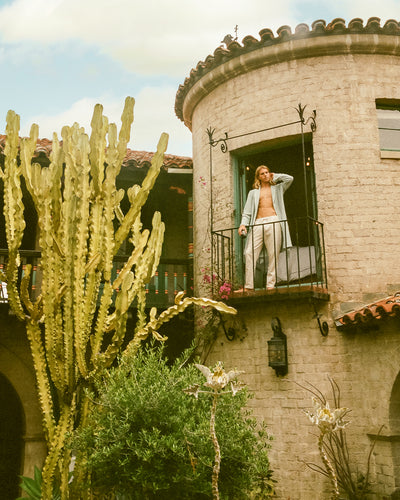 Wearing the Dandy Del Mar Tropez Terry Cloth Robe in Gulf Blue, a person stands on a stone balcony with a round tower, surrounded by tall cacti and greenery beneath a partly cloudy sky.