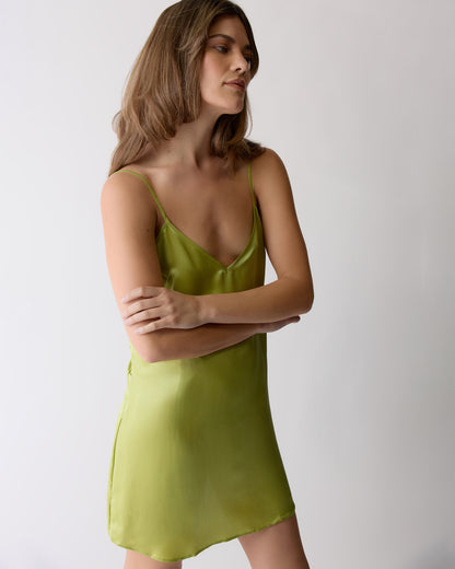 A woman poses against a plain background in The Adonia Satin Slip Dress in lime by Dandy Del Mar, featuring thin straps and a mini silhouette, arms crossed and gazing to the side.