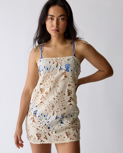 A woman in the Dandy Del Mar Mara Tie Strap Dress - Scallop stands against a plain light background, featuring beige scalloped crochet over blue undergarments.