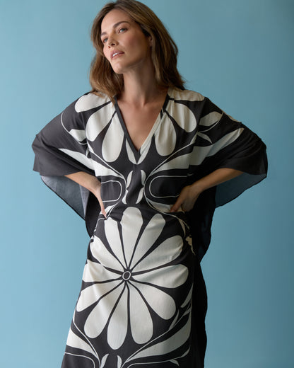 A woman models The Tropea Kaftan - Albatross by Dandy Del Mar, featuring a bold black and white floral print, while standing against a plain light blue background and looking slightly upward.