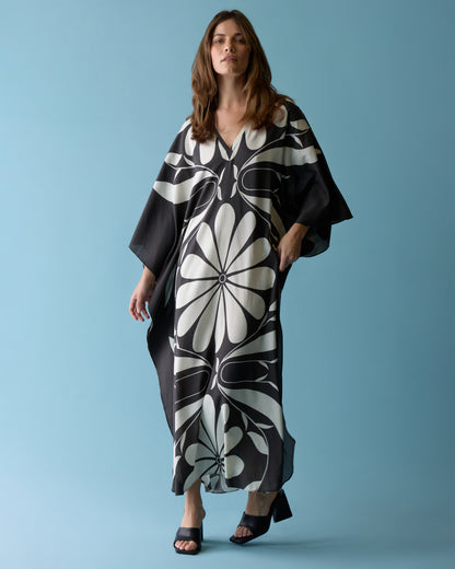 A woman stands against a blue background wearing Dandy Del Mar's The Tropea Kaftan - Albatross and black heeled sandals.
