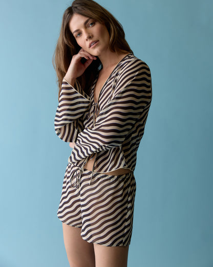 A woman poses against a blue backdrop, wearing Dandy Del Mar's The Curacao Short - Tierra, paired with a matching striped long-sleeve top and an elastic waistband, as she touches her hair.