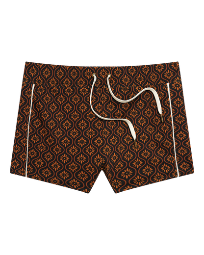 The Cassis Square Cut Swim Brief - Cacao by Dandy Del Mar: men's swim trunks crafted from recycled polyester with a brown, orange, and black geometric floral design and white drawstrings, shown against a black background.
