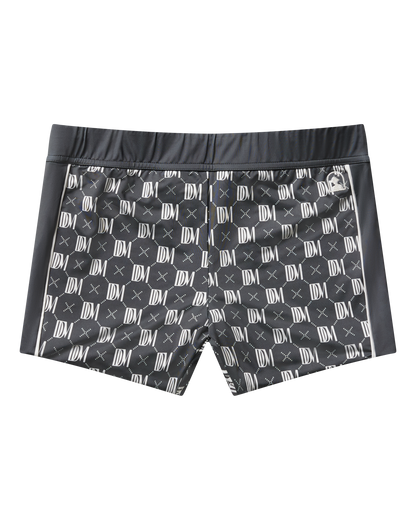 The Cassis Square Cut Swim Brief - DDM Monogram by Dandy Del Mar features a white monogram pattern on black recycled polyester, with a modern European fit, fitted waistband, and side panels—shown against a plain black background.