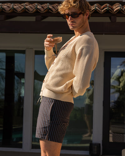 A person in sunglasses, a beige cardigan, and Dandy Del Mar’s The Ventura Volley Shorts - Moontide stands outdoors holding a glass, with building and palm tree reflections behind them.