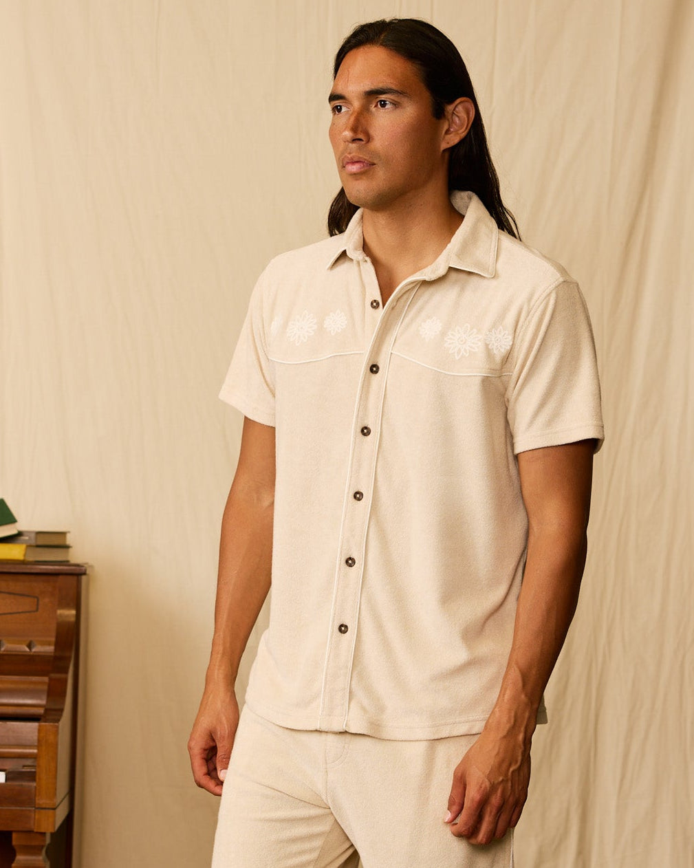 A man with long hair stands indoors wearing the Dandy Del Mar Gaucho Terry Cloth Shirt in beige and matching shorts. A piano and books are partially visible on the left.
