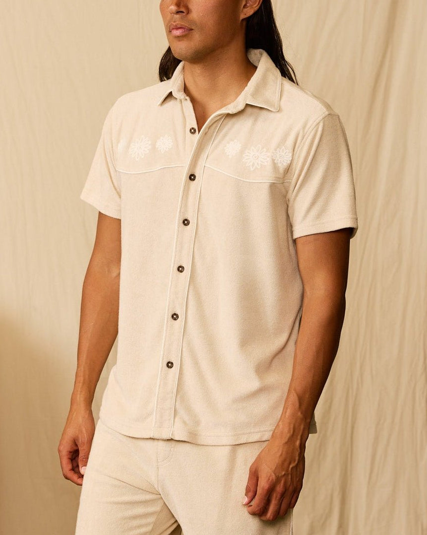 A man with long hair stands indoors wearing the Dandy Del Mar Gaucho Terry Cloth Shirt in beige and matching shorts. A piano and books are partially visible on the left.