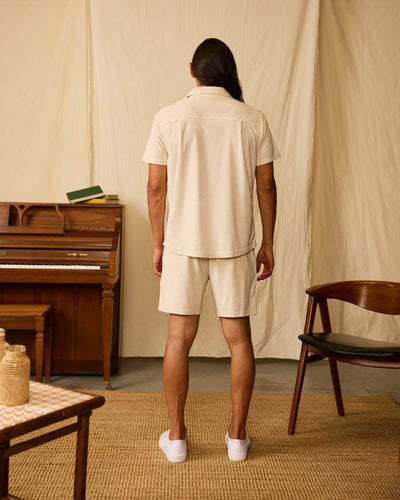 A person with long hair stands facing away, wearing the Dandy Del Mar Gaucho Terry Cloth Shirt in Alabaster and shorts near a piano, chair, and table in a neutral-toned room.