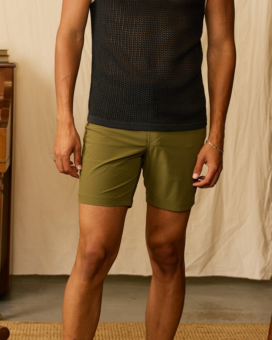 Indoors, a person wears a black mesh tank top, flip-flops, and Dandy Del Mar’s The Cavoli Swim Shorts - Berdea. In the background are a wooden chair, piano, table, and books.