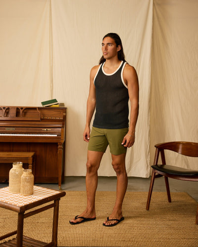 A person stands indoors on a woven rug, wearing a black tank top, Dandy Del Mar’s The Cavoli Swim Shorts - Berdea, and flip-flops. In the background are a piano, wooden chair, and table with decor items.
