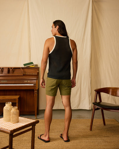 A man stands indoors with his back to the camera, wearing a black sleeveless top, flip-flops, and Dandy Del Mar's The Cavoli Swim Shorts - Berdea. A piano, chair, and table can be seen in the background.
