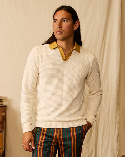 A man with long dark hair stands with one hand in his pocket against a beige backdrop, wearing the Dandy Del Mar Sebastian LS Polo Shirt in Vintage Ivory over a gold collared shirt and striped pants.