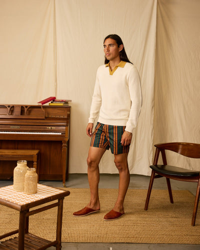 A man stands indoors wearing the Dandy Del Mar Sebastian LS Polo Shirt in Vintage Ivory over a yellow collared shirt, paired with striped shorts and brown slippers. A piano, books, chair, and table are visible in the background.