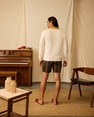 A person with long hair stands facing away, wearing the Dandy Del Mar Sebastian LS Polo Shirt in Vintage Ivory and striped shorts in a room with a piano, wooden chair, and small table.