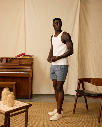 A man in a white tank top, Dandy Del Mar's The Rhodes Twill Shorts in Abyss, and white sneakers stands in a room with a piano, chair, and small table against a beige backdrop.