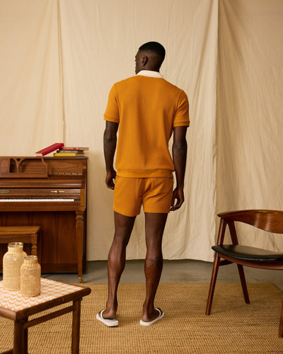 A man in the Dandy Del Mar Cannes Waffle Knit Polo Shirt in Burnt Sienna faces away in a room with a piano, chair, table, and neutral drapes.