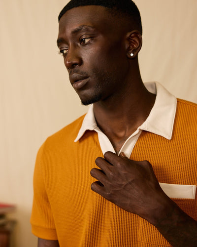 A man wearing the Dandy Del Mar Cannes Waffle Knit Polo Shirt in Burnt Sienna, featuring a white collar and pocket detail, looks to the side while lightly holding his shirt.