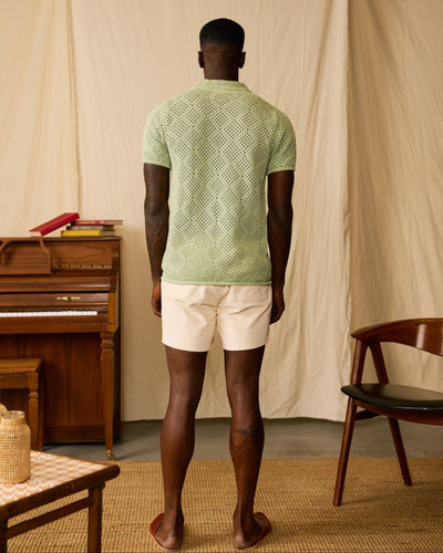 A man faces away, wearing the Dandy Del Mar Antibes Crochet Polo Shirt in Pistachio and cream shorts, in a room with a piano, books, a chair, and a woven rug.