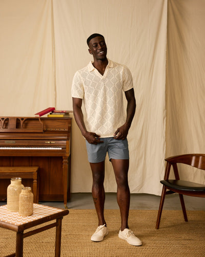 Indoors, a man wears the Dandy Del Mar Antibes Crochet Polo Shirt in Vintage Ivory, paired with blue shorts and cream sneakers. Behind him are a piano, books, a chair, and a table with baskets.