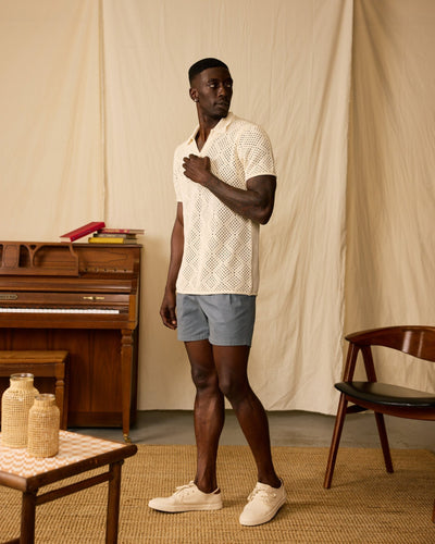 A man stands indoors wearing the Dandy Del Mar Antibes Crochet Polo Shirt in Vintage Ivory, paired with gray shorts and cream sneakers. Behind him are a piano, chair, and small table on a woven rug.