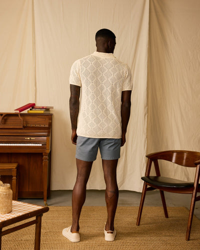 A man faces away in a room with a piano, chair, and beige drapes, wearing the Dandy Del Mar Antibes Crochet Polo Shirt in Vintage Ivory along with blue shorts and white shoes.