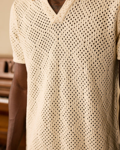 Someone wearing The Antibes Crochet Polo Shirt in Vintage Ivory by Dandy Del Mar, a beige short-sleeved V-neck knit shirt with a subtle diamond pattern, shown from shoulders to waist.