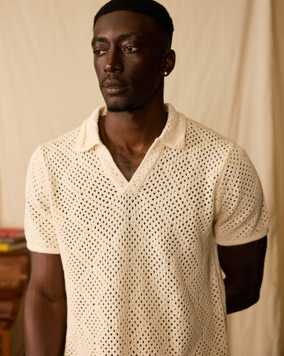 A man wearing The Antibes Crochet Polo Shirt in Vintage Ivory by Dandy Del Mar stands against a plain beige background, looking slightly to the side.