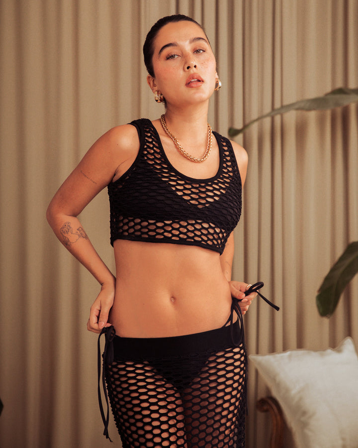 Person standing indoors wearing a black fishnet bikini top and bottoms with a curtain backdrop, complemented by **The Montserrat Tank - Onyx** from **Dandy Del Mar**. A potted plant is partially visible in the scene.