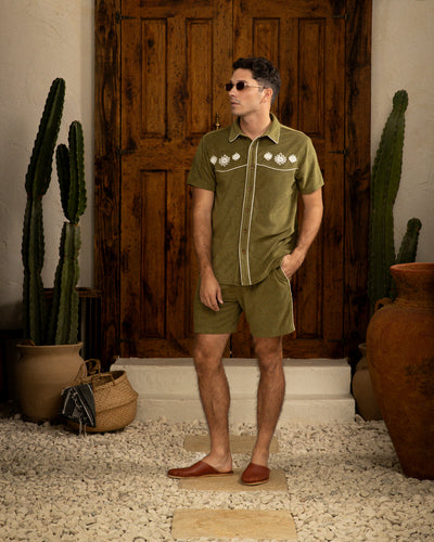 A man in an olive green short-sleeve shirt and Dandy Del Mar's The Gaucho Terry Cloth Shorts - Arbequina stands before a wooden door, surrounded by potted cacti and clay pots on a stone floor.