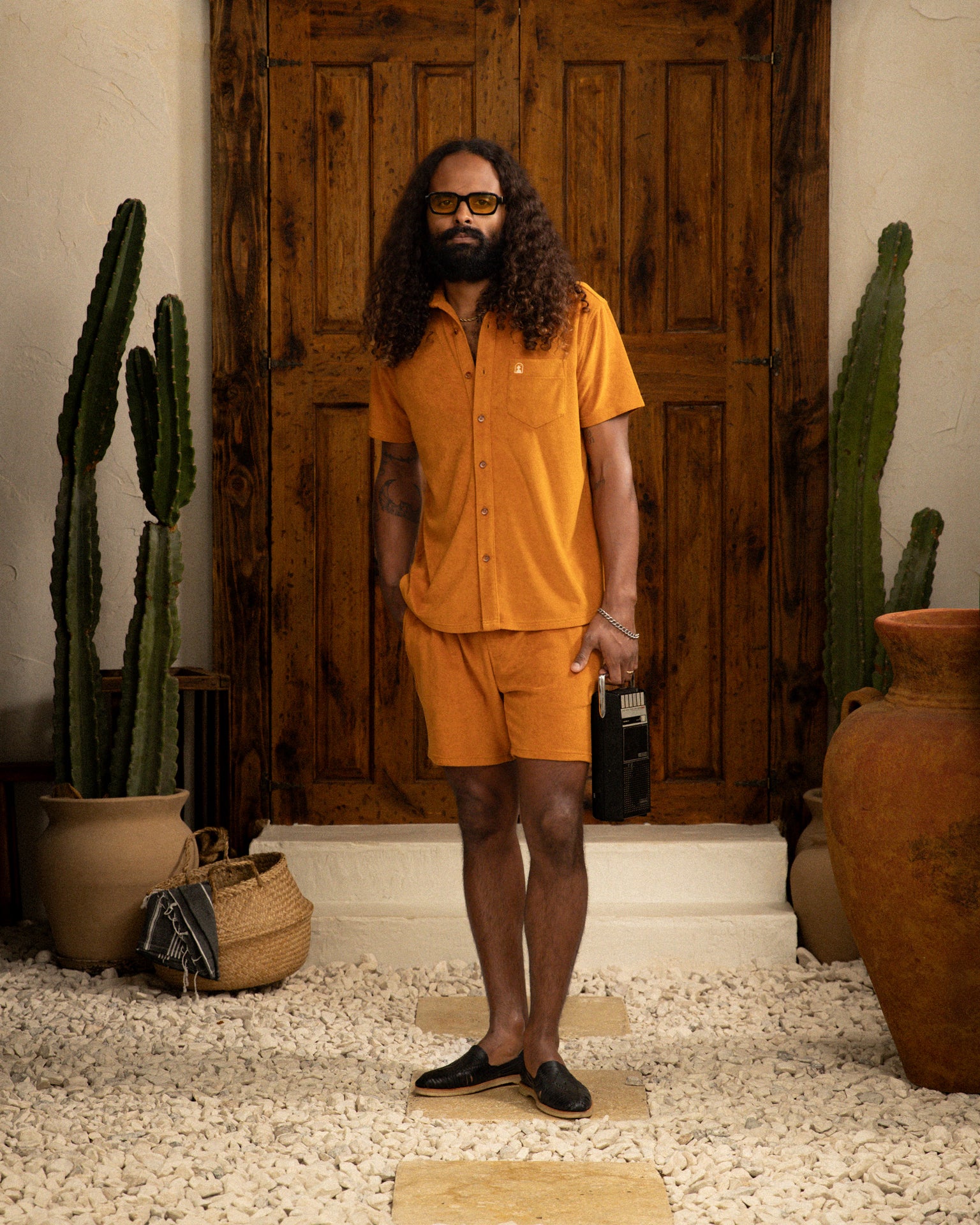 A person with long curly hair and a beard stands in front of a wooden door, wearing an orange Tropez Shirt by Dandy Del Mar, crafted from terry cloth. They hold a small black bag amidst cacti and large pots that enhance the serene Côte d'Azur-inspired scene. The shirt is called The Tropez Shirt - Burnt Sienna.