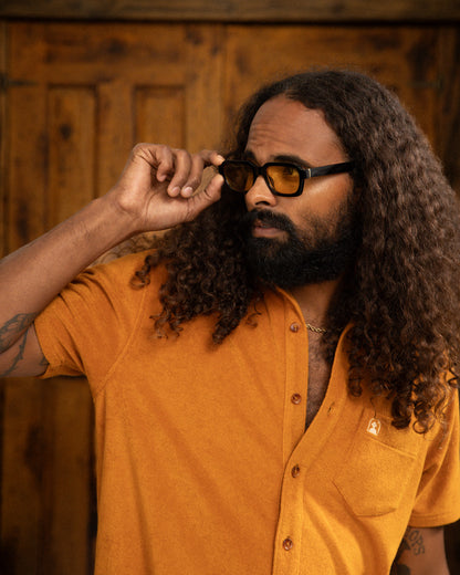 A person with long curly hair and sunglasses stands against a wooden backdrop, exuding effortless Côte d'Azur charm while wearing the Burnt Sienna Tropez Shirt by Dandy Del Mar.