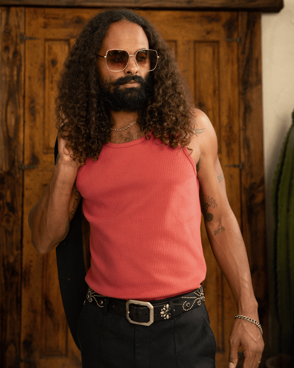 A man with long curly hair and a beard, wearing a Dandy Del Mar Milan Rib Tank in Currant with dropped armholes, aviator sunglasses, and a belt with a large buckle stands confidently in a room with wooden floors.