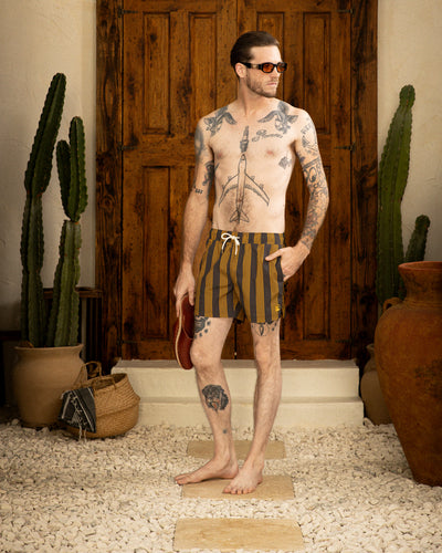 A tattooed man stands barefoot on a stone path, wearing sunglasses and Dandy Del Mar's The Ventura Volley Shorts - Albatross, holding flip-flops, with potted plants and a wooden door in the background.