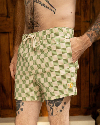A person wearing The Ventura Volley Shorts - Arbequina by Dandy Del Mar, featuring a green and white checkered pattern, drawstring, and elastic waist. Visible torso and leg tattoos; standing indoors.