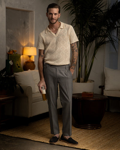 A man stands in a living room holding a drink, wearing the Dandy Del Mar Antibes Crochet Polo Shirt in Vintage Ivory, with gray trousers and dark loafers. A sofa, lamp, and large plant are seen in the background.