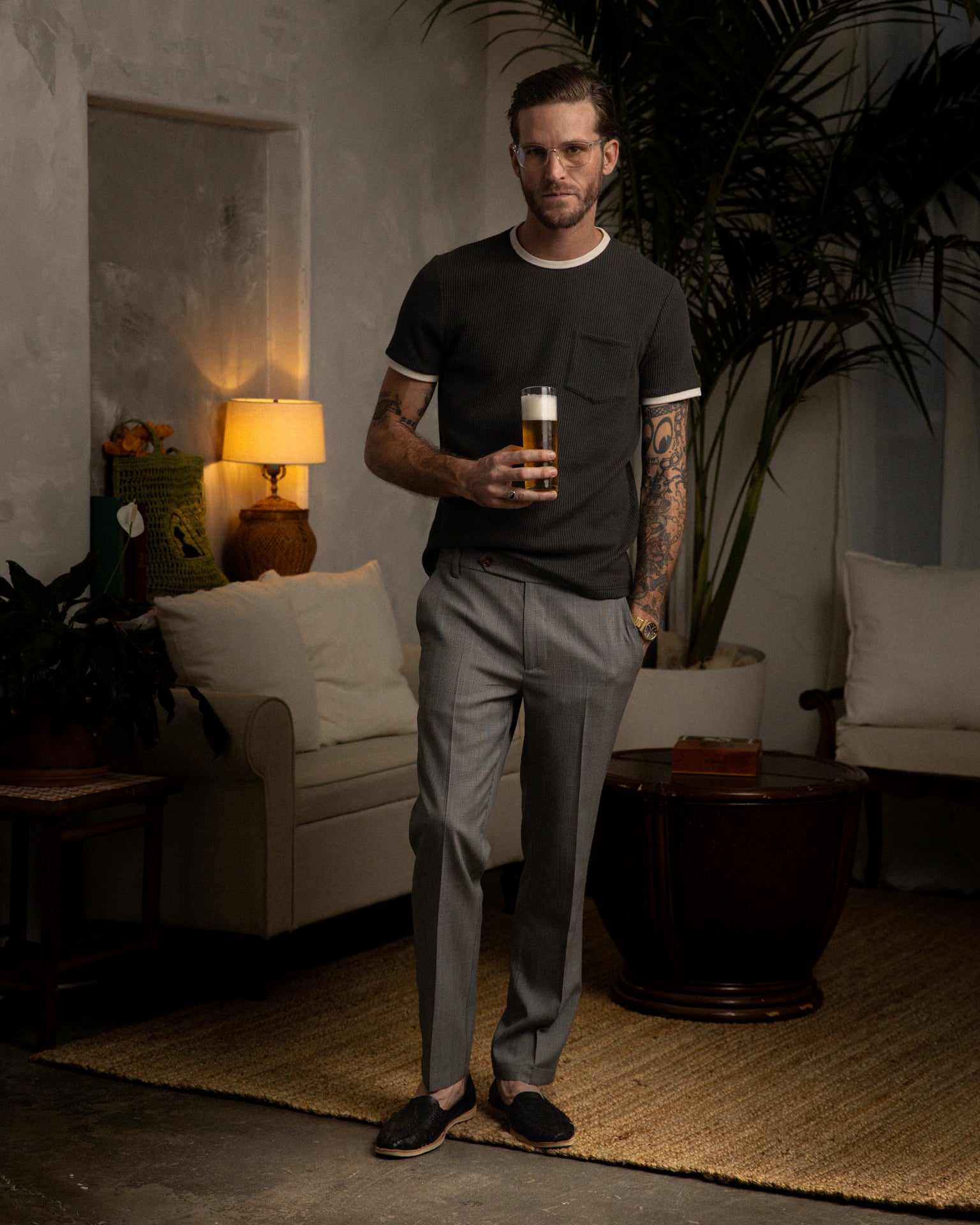 A tattooed man stands in a living room with a sofa, lamp, and plants, holding a drink. He's comfortably dressed in the dark Albatross Cannes Waffle Knit Tee by Dandy Del Mar and gray pants.