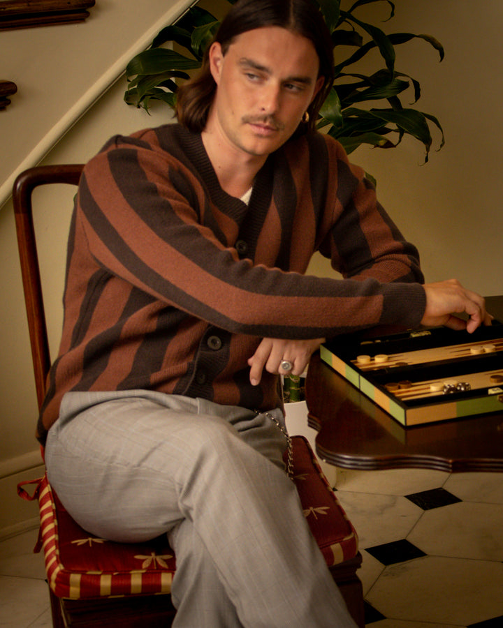 A person with long hair sits on a striped chair, wearing The Florence Knit Cardigan - Garam by Dandy Del Mar with ribbed trims and light pants, next to an open backgammon set on a wooden table.