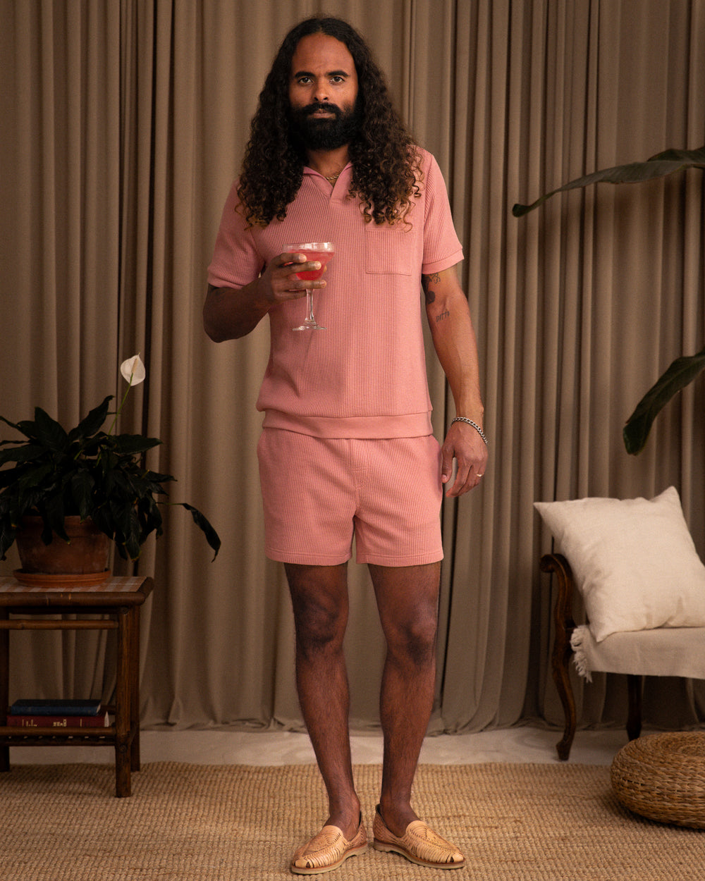 A man with long curly hair stands holding a cocktail, wearing a pink short-sleeve shirt, matching Dandy Del Mar Cannes Waffle Knit shorts in Spanish Rose, and tan woven shoes in a softly lit room with plants and neutral decor.