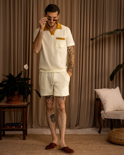 A man wears the Dandy Del Mar Cannes Waffle Knit Polo Shirt in Vintage Ivory and shorts with brown accents indoors, lifting his sunglasses. A potted plant, side table, chair, and beige curtains are in the background.
