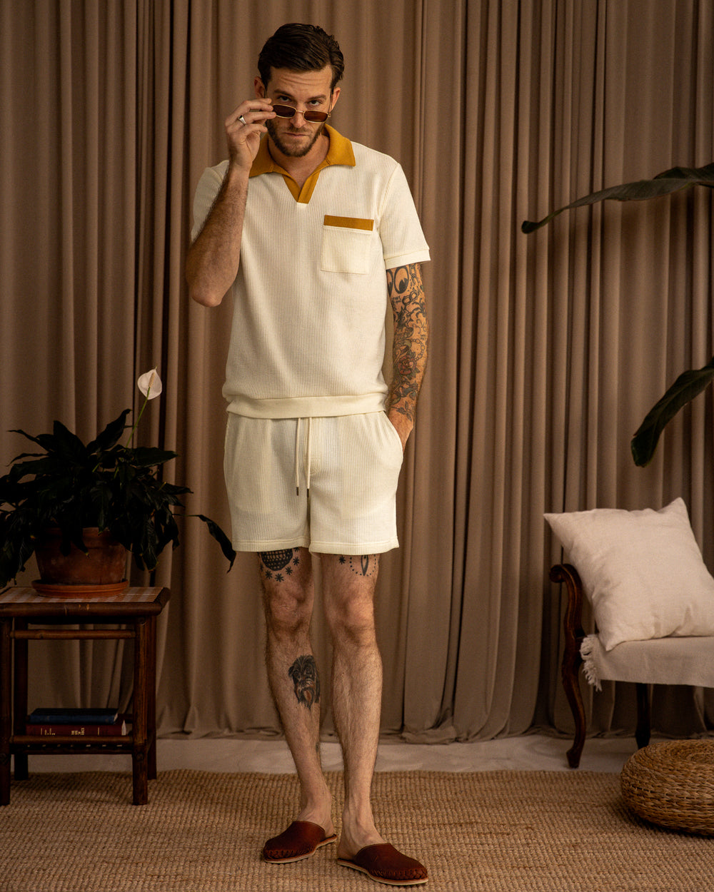 Man in a casual white and yellow polo shirt made of 100% cotton and Dandy Del Mar Cannes Waffle Knit Shorts - Vintage Ivory, standing and talking on a cellphone in a room with neutral tones.