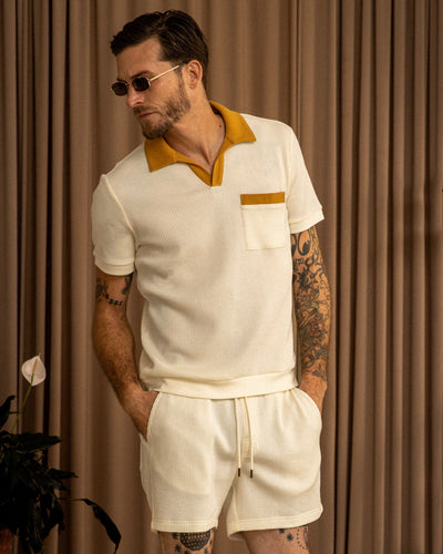 A man in the Dandy Del Mar Cannes Waffle Knit Polo Shirt in Vintage Ivory, featuring a mustard collar and matching shorts, stands in front of beige curtains with his hands in his pockets.