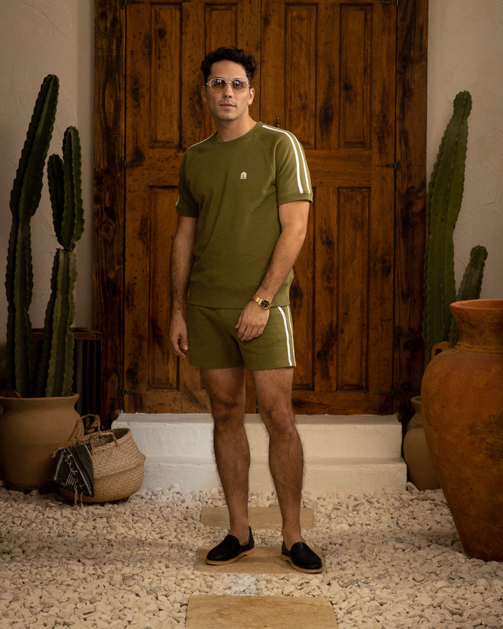 A man in a green Dandy Del Mar Marseille Pullover - Arbequina t-shirt and shorts standing in front of a cactus.
